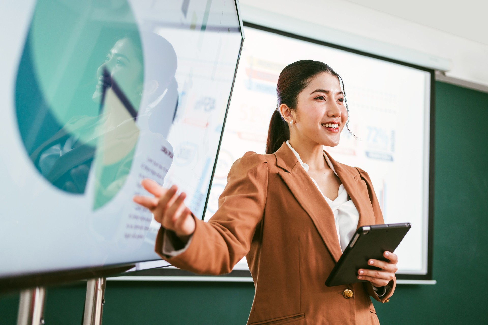 A professional Asian business woman presents data on screens in a board meeting room, engaging her audience with confidence and clarity. A businessman wearing a blazer while using a tablet for meeting